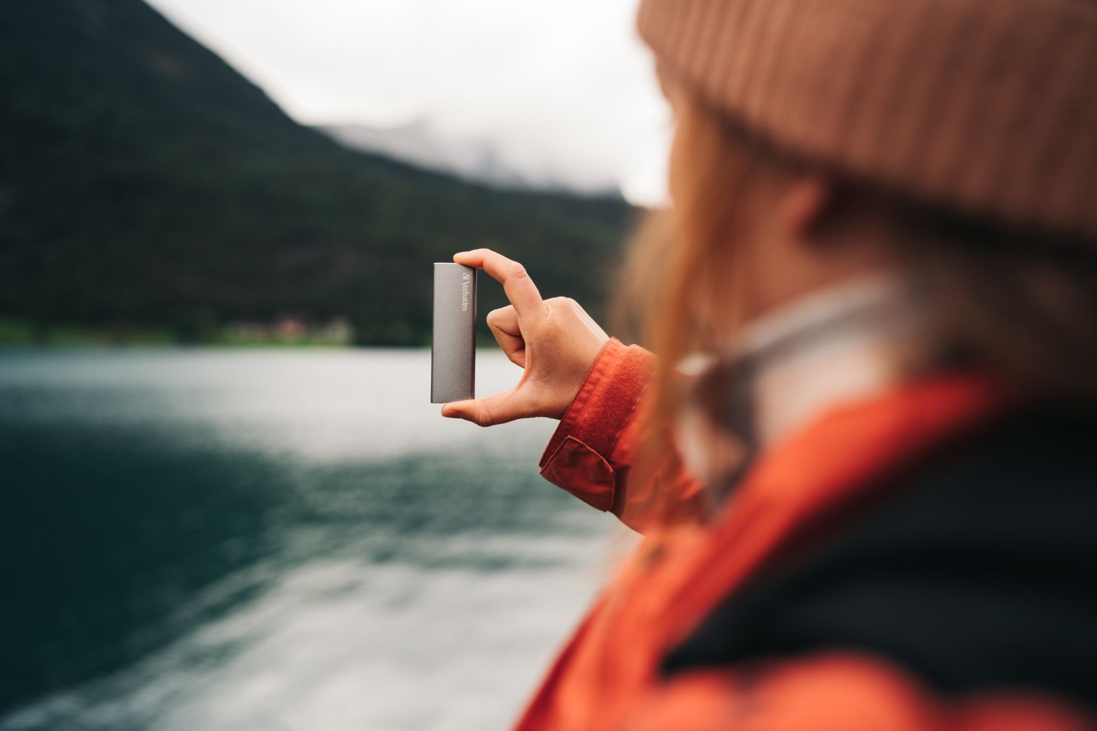 Osoba trzyma szary prostokąt, jezioro i góry w tle.