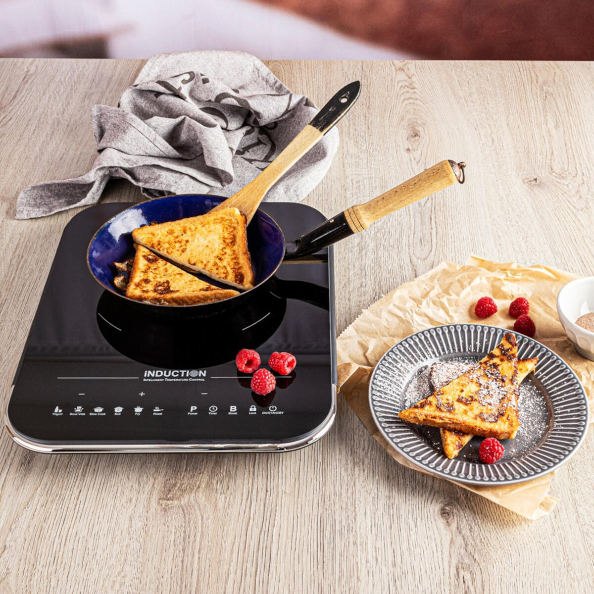 Tosty francuskie w niebieskiej patelni na płycie indukcyjnej, z malinami i talerzem tostów z cukrem.