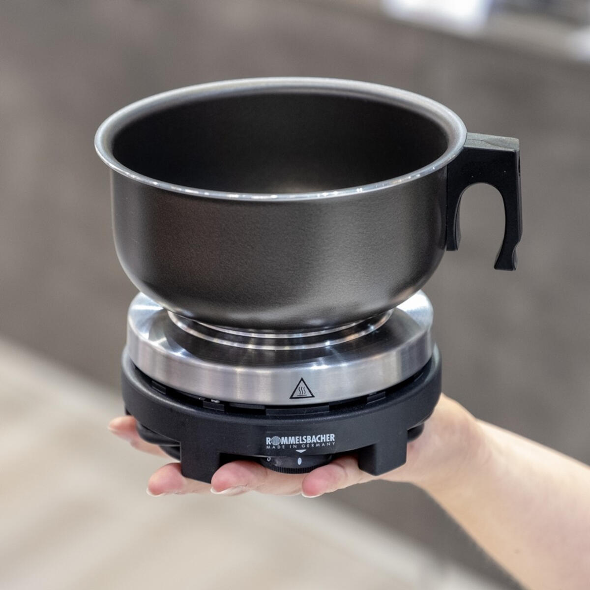Un caquelon à fondue noir sur un support noir et argent, tenu par une main.