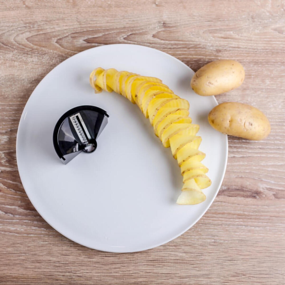 Aardappelspiraalsnijder met aardappelen op wit bord en houten achtergrond.