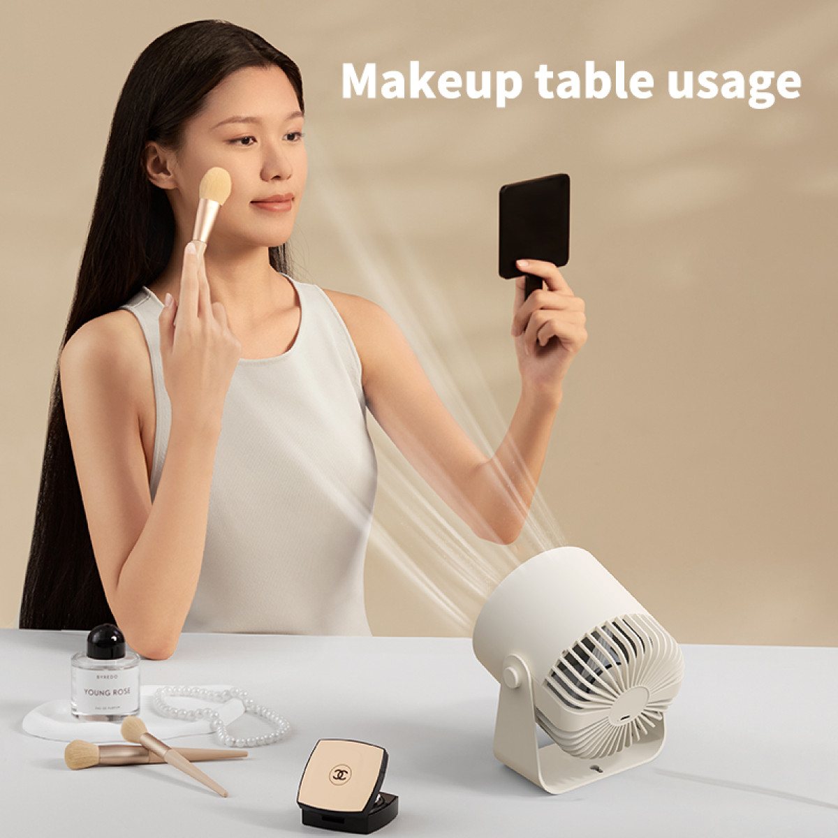 Woman applying makeup while a small fan blows air. Makeup and brushes on the table.