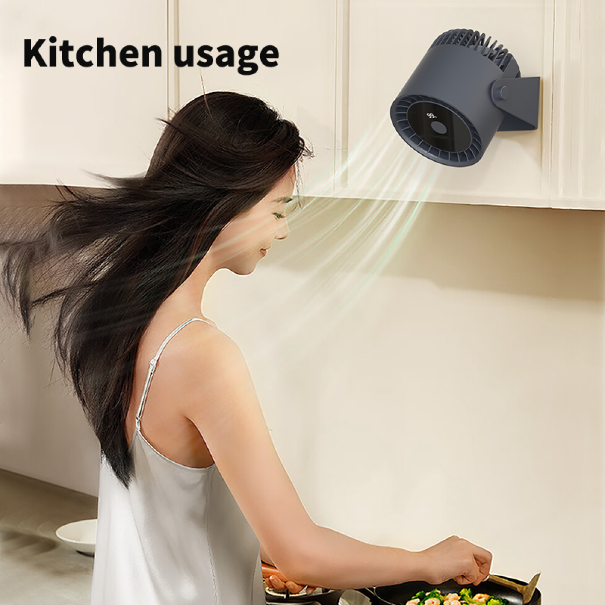 A woman in a kitchen with a fan mounted on the wall, directing air towards a pan.