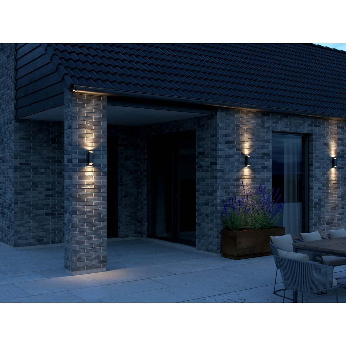 Una casa moderna con tres luces de pared. Las luces brillan sobre una pared de ladrillo y una jardinera de madera.