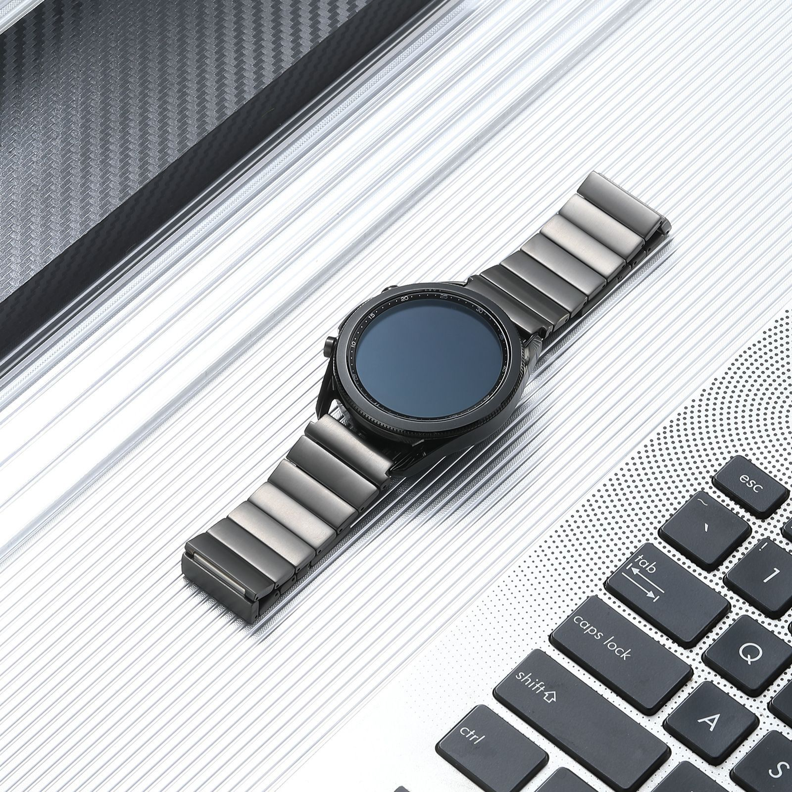 Eine schwarze Smartwatch mit Metallarmband liegt auf einer metallischen Oberfläche neben einer Laptop-Tastatur.