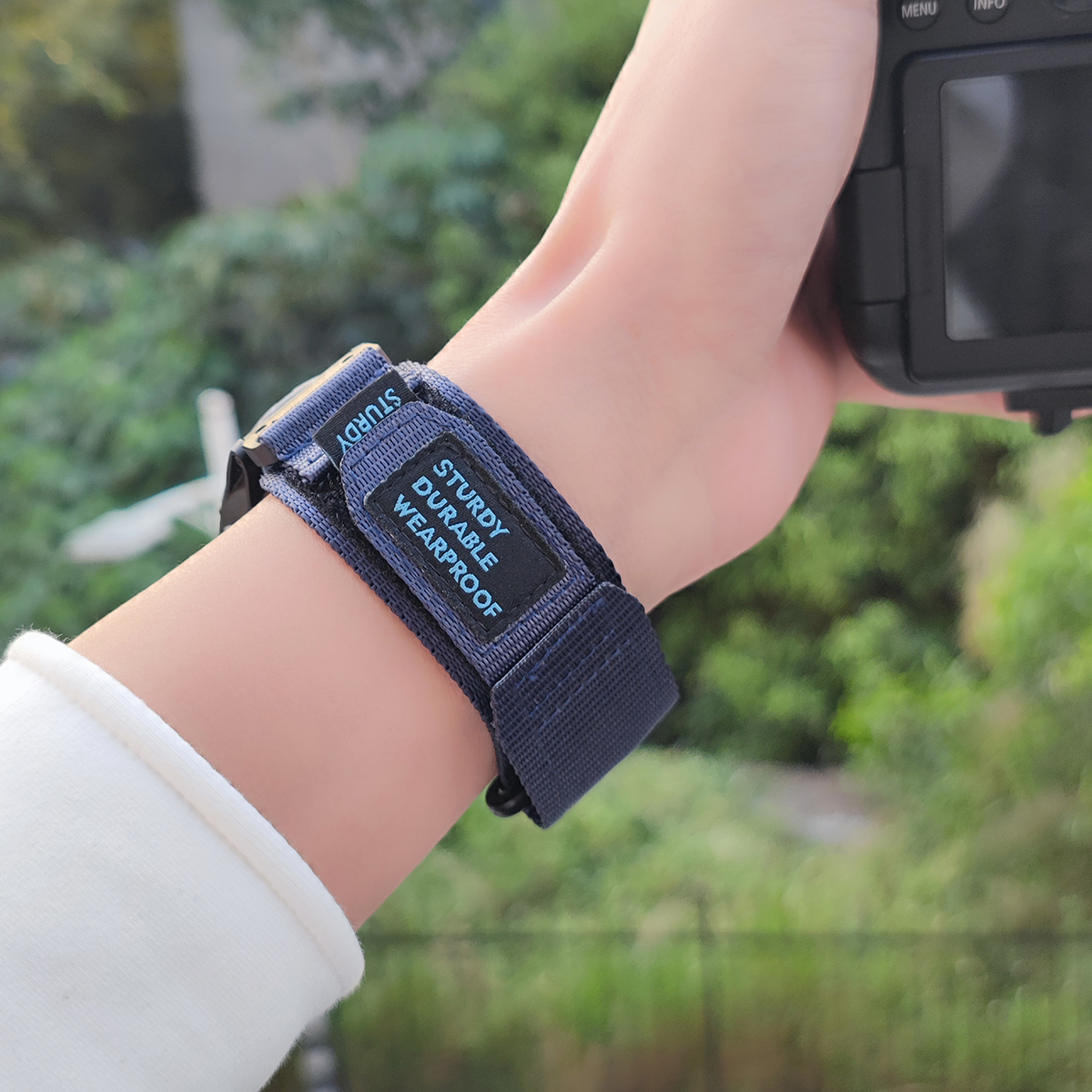 Hand mit blauem Kameragurt, Text: Sturdy Durable Wearproof. Kamera im Hintergrund.