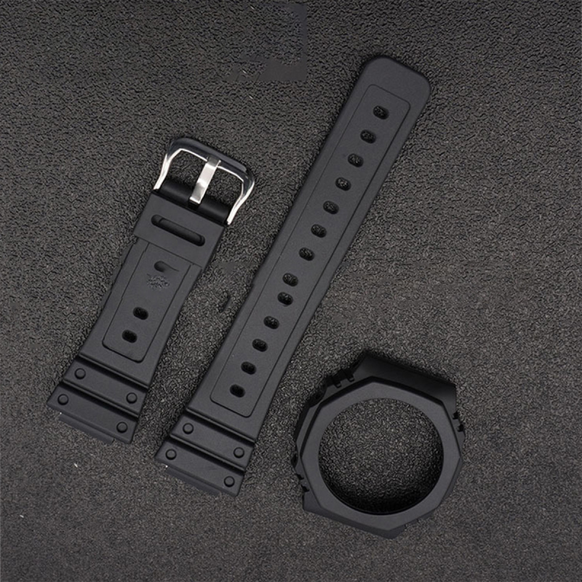 Schwarze Uhrenteile auf dunkler Oberfläche: Uhrenarmband, Schnalle und Uhrenlünette.