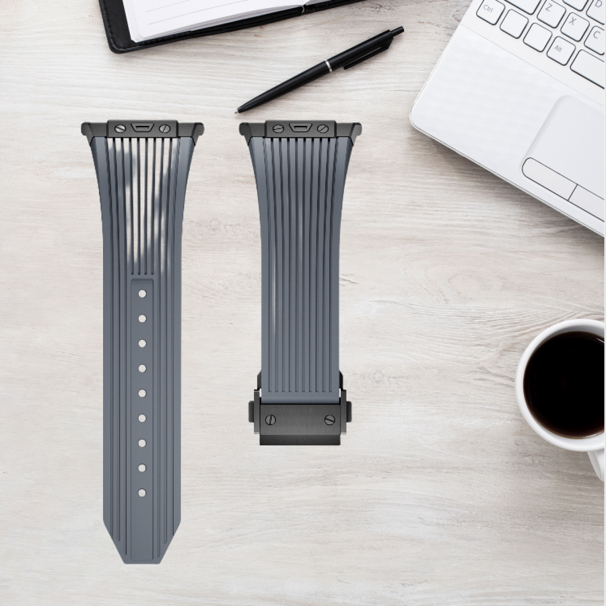 Due cinturini grigi per orologi su una superficie di legno con una penna, un taccuino e una tazza di caffè.