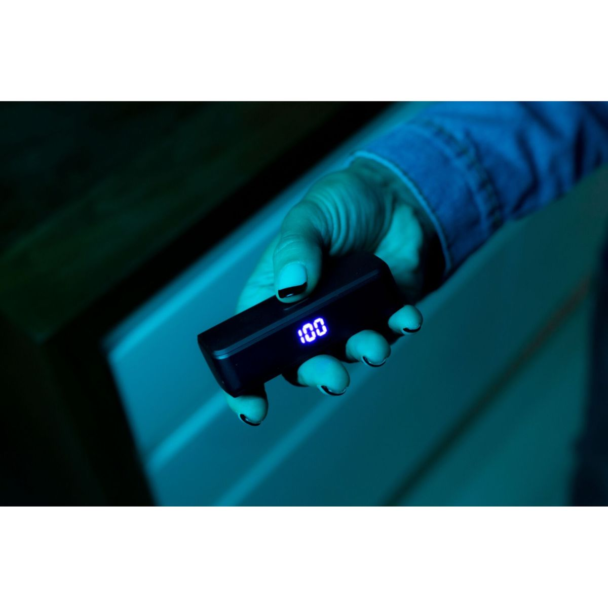 Mano che tiene un dispositivo nero con un display blu brillante che mostra 100. Lo sfondo è blu-verde scuro.