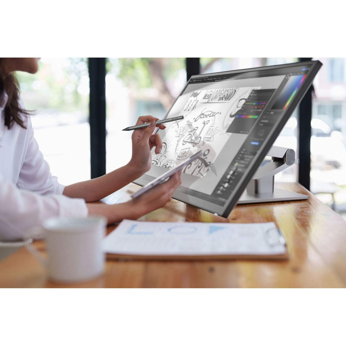 Kobieta używa pióra i rysika na dużym monitorze cyfrowym. Na biurku stoi kubek.