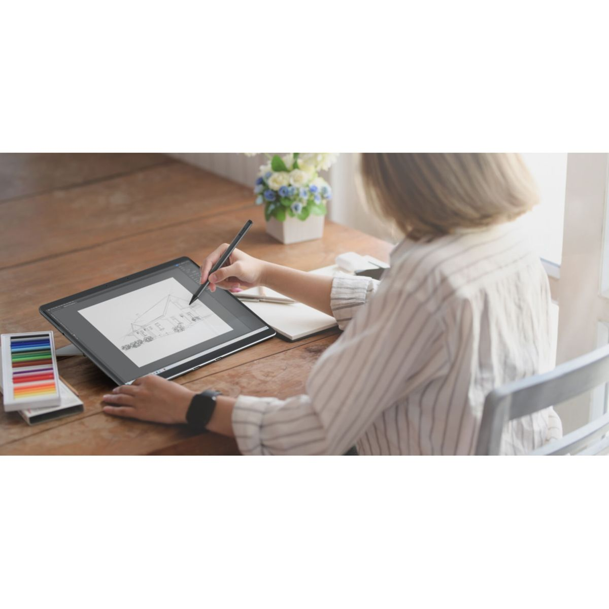 Osoba rysuje na tablecie rysikiem. Kwiaty i notatnik na biurku.