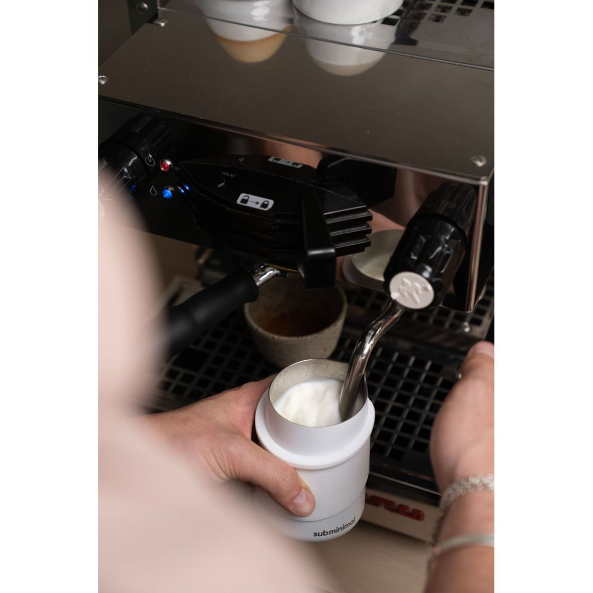 Eine Person schäumt Milch für einen Latte mit einer Espressomaschine auf, mit einer weißen Tasse in der Hand.