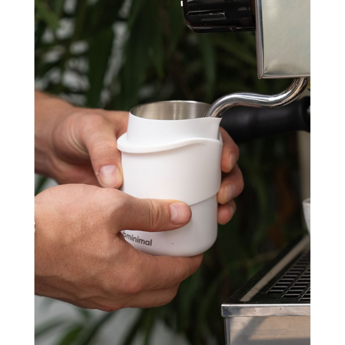 Hände halten einen weißen Milchaufschäumkrug neben einer Espressomaschine.