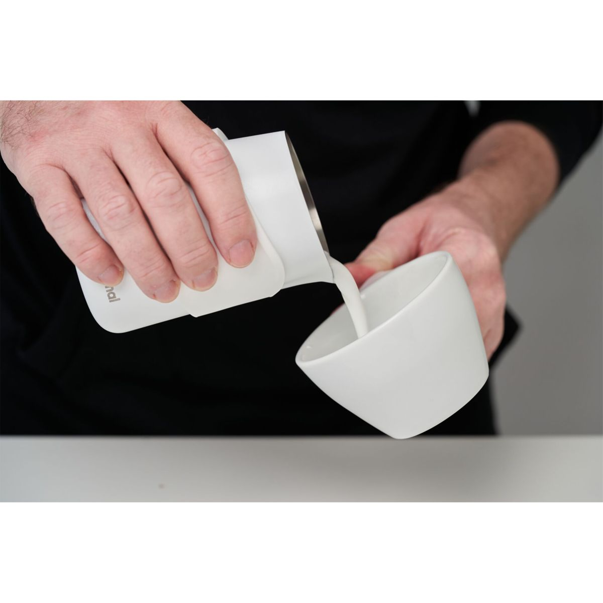 Hände gießen Milch aus einem weißen Krug in eine weiße Schüssel. Die Szene spielt sich auf einer weißen Oberfläche ab.