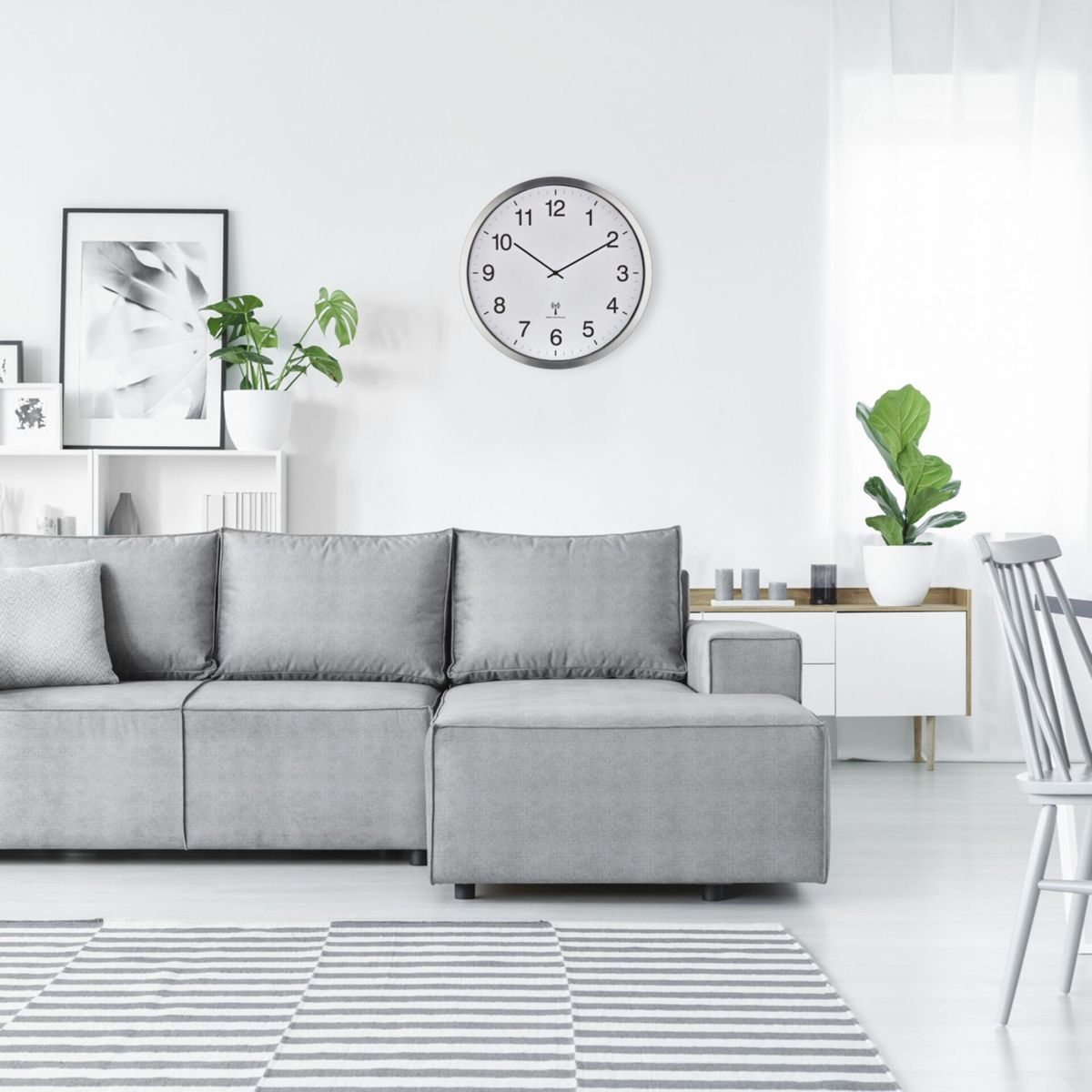 Wohnzimmer mit grauem Sofa, Teppich, Wanduhr und Pflanzen. Weiße Wände und Holzboden.