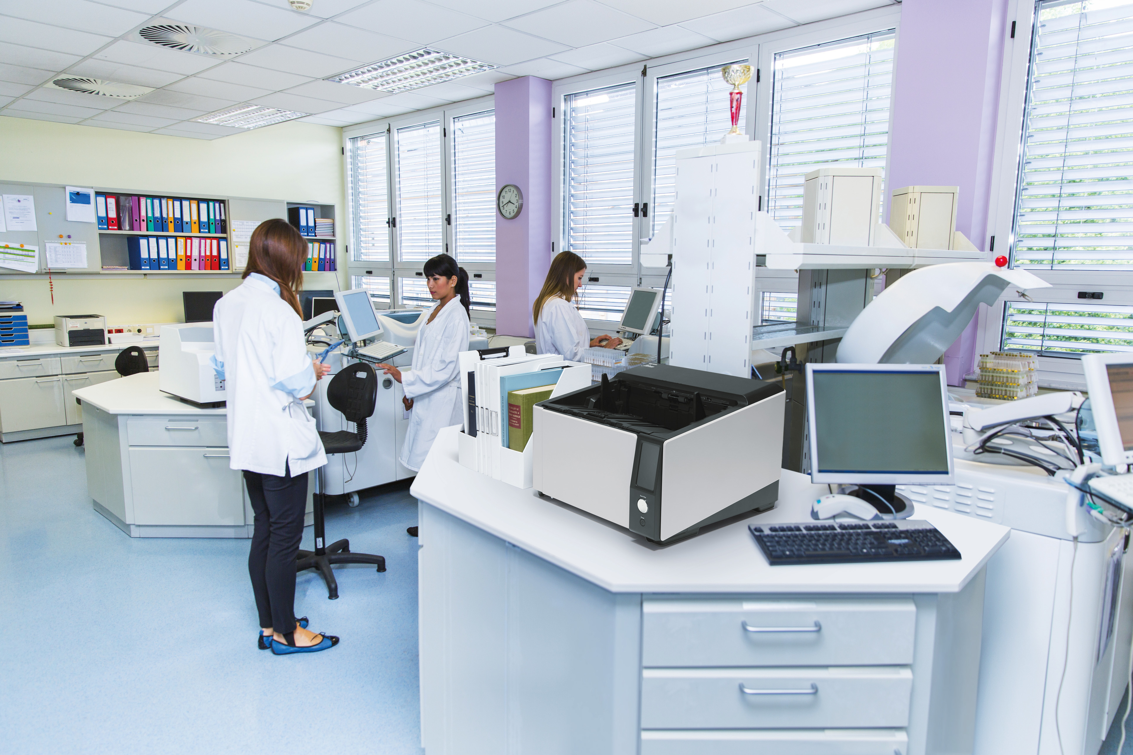 Laboratoriumscène met drie mensen in laboratoriumjassen. Apparatuur en een printer zijn zichtbaar.
