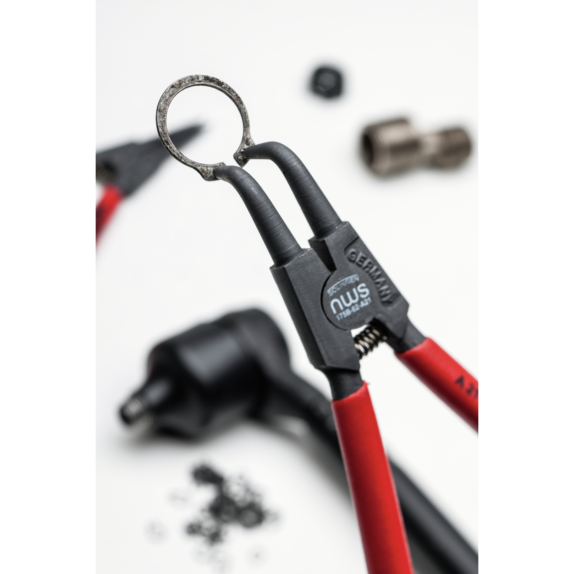 Nahaufnahme einer Ringzange mit roten Griffen, die einen Metallring greift, mehrere Werkzeuge und Teile sind unscharf.