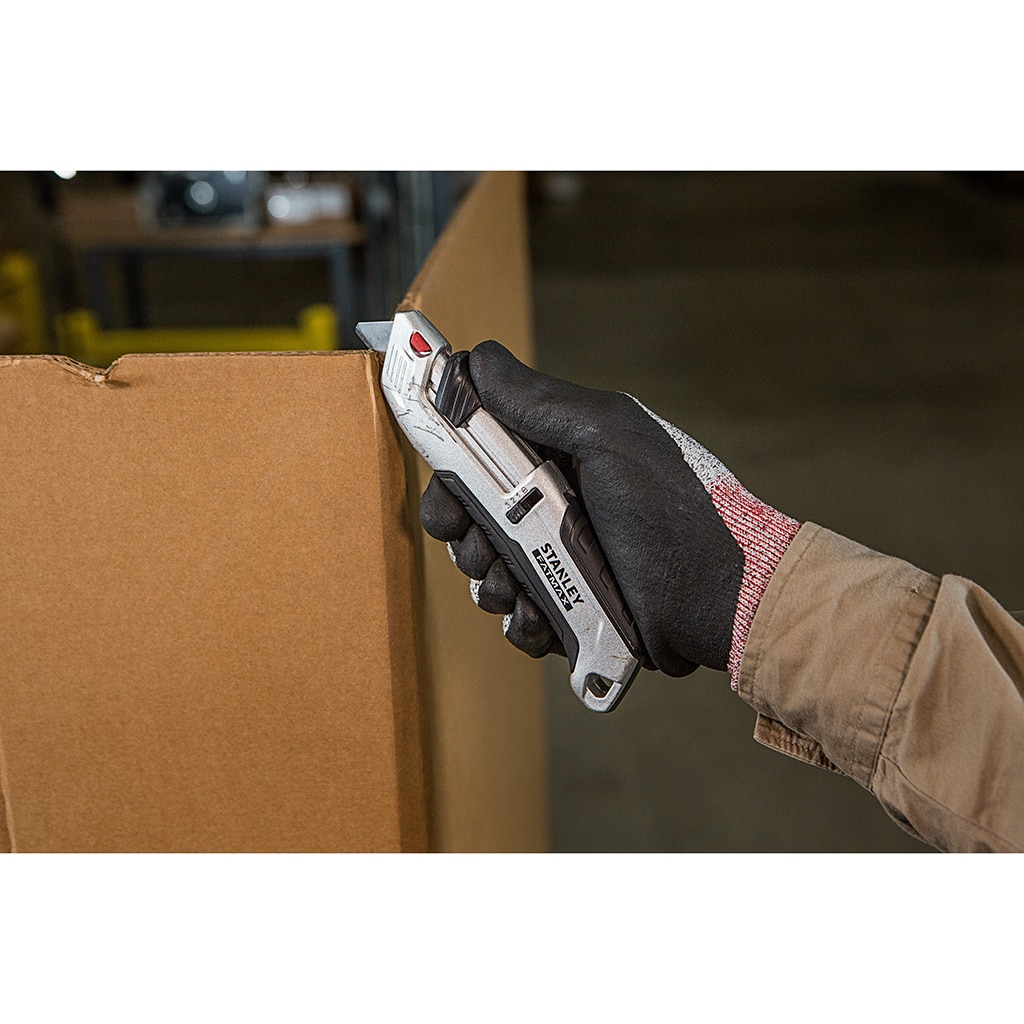 Eine behandschuhte Hand schneidet mit einem silbernen Universalmesser Pappe. Kisten sind im Hintergrund sichtbar.