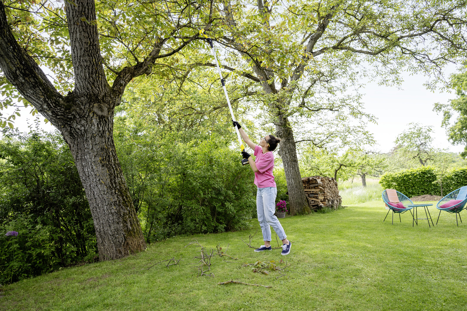 Kobieta przycina gałęzie drzew z długim trzonkiem w słonecznym ogrodzie.