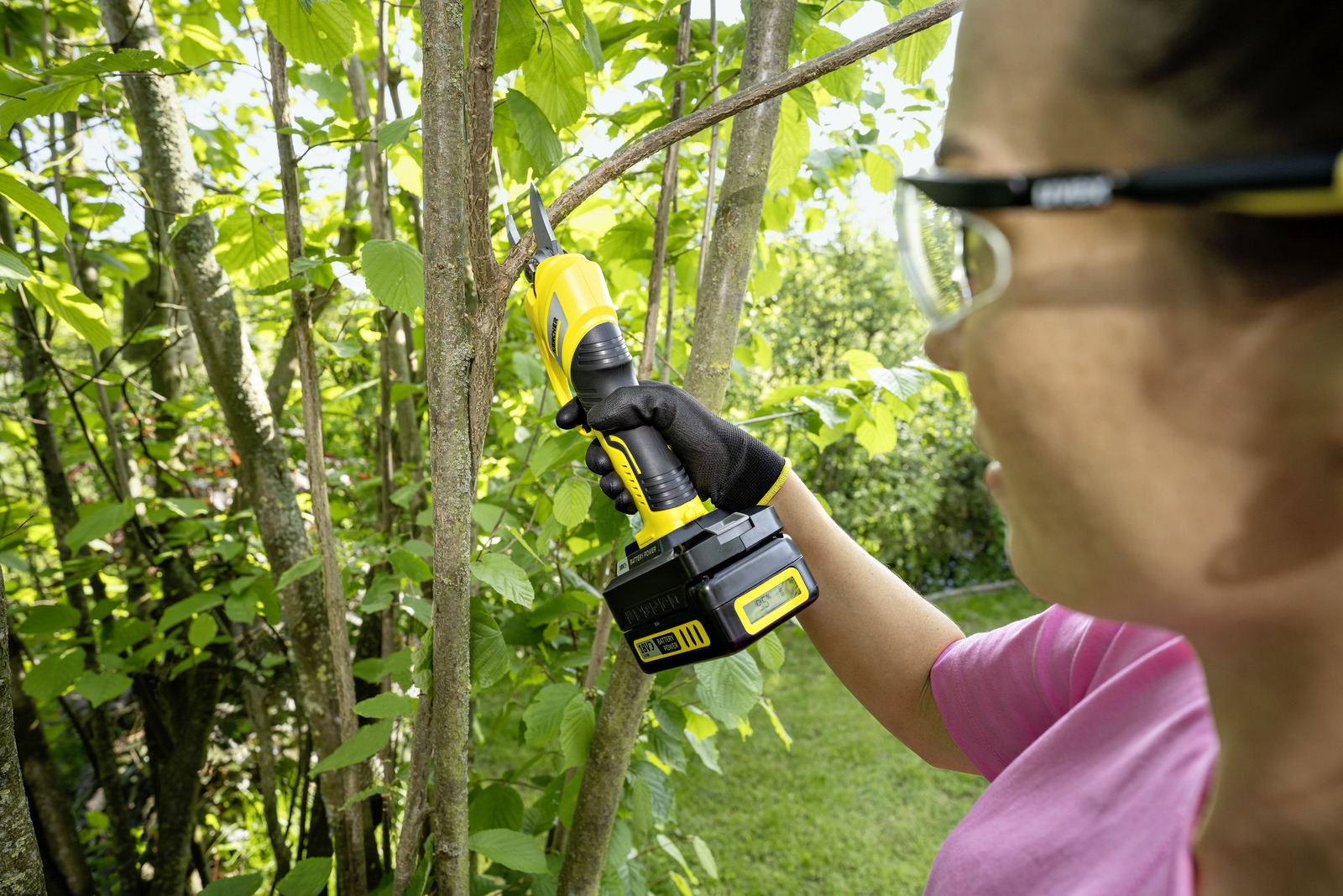 Kobieta używa żółto-czarnej piły do przycinania gałęzi. Scena rozgrywa się na zewnątrz, z zielonymi drzewami.