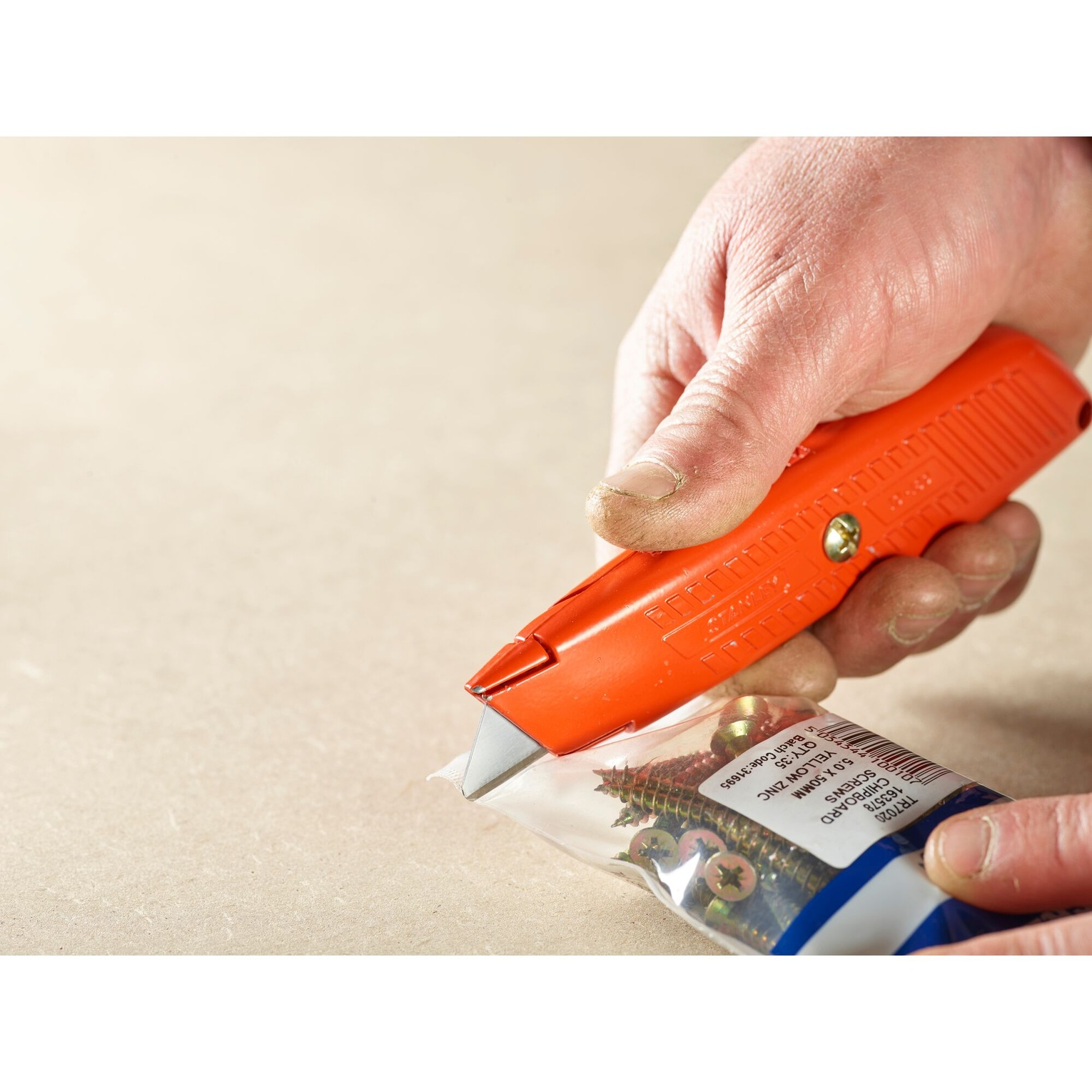 Una mano sosteniendo un cuchillo utilitario naranja, cortando una bolsa de plástico con tornillos.