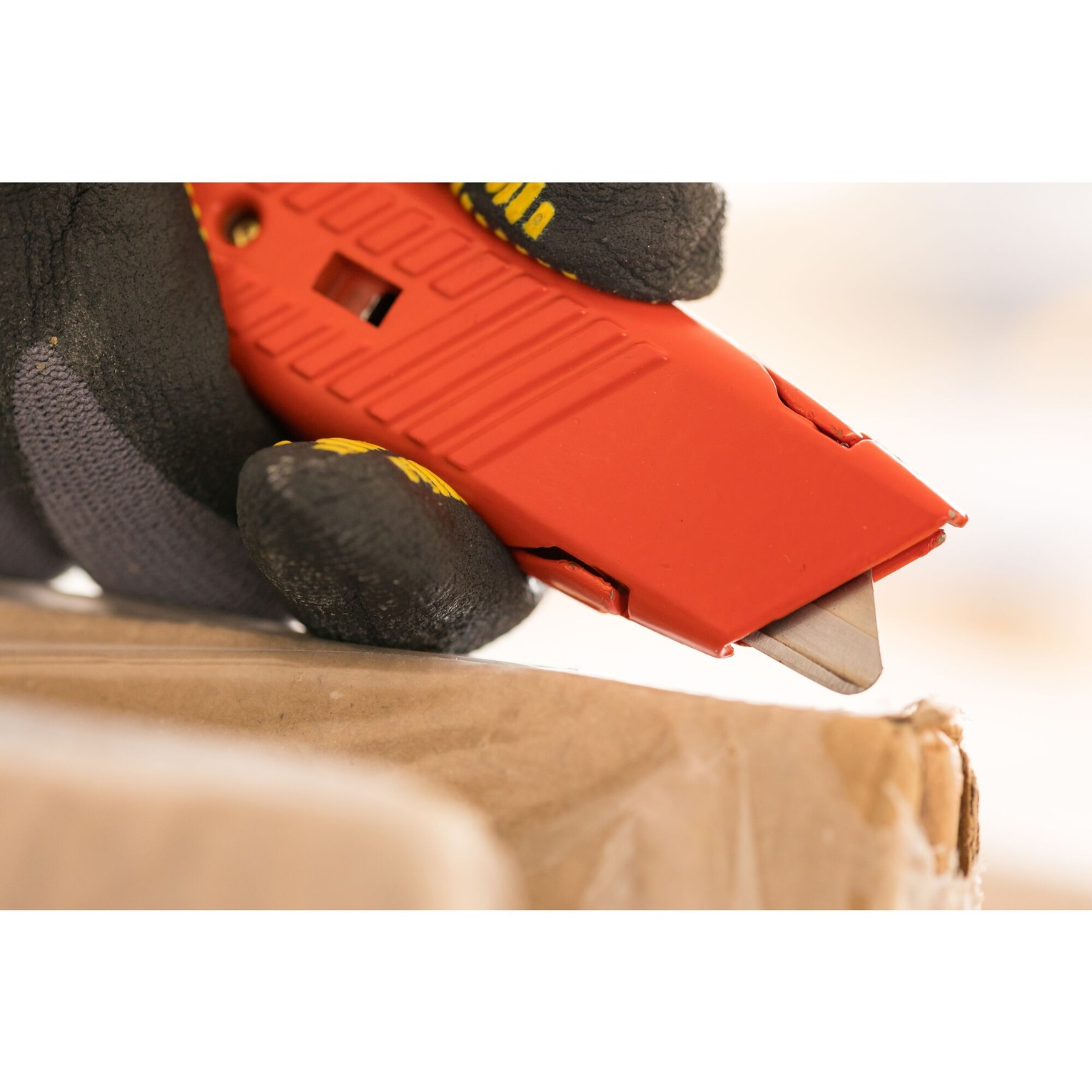Una mano con un guante negro sostiene un cuchillo rojo cortando cartón.