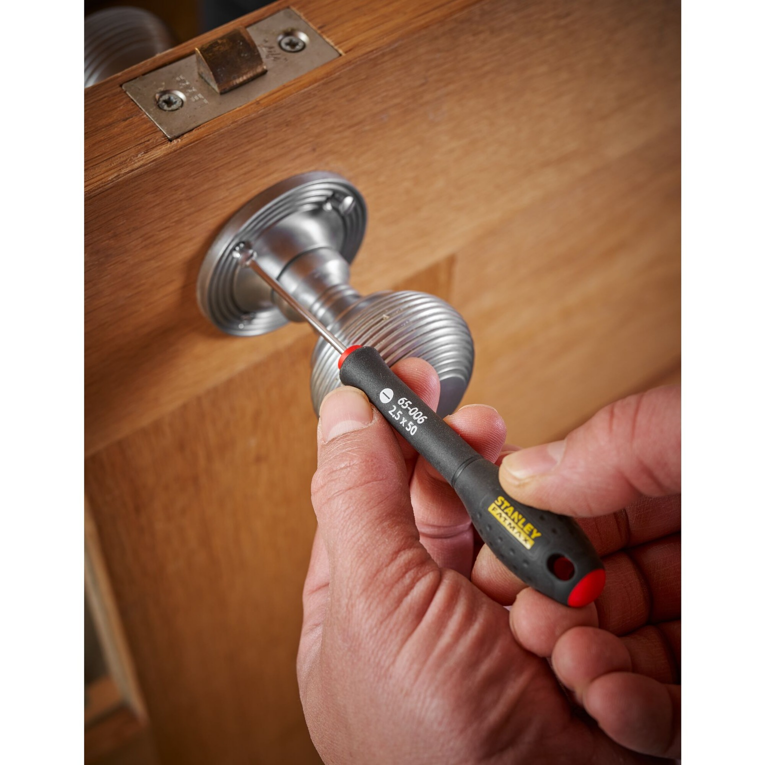 Ręka używa śrubokręta do dokręcania śruby w klamce. Narzędzie jest czarne i czerwone.