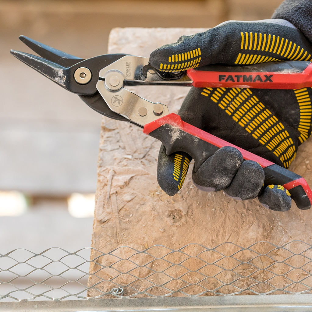 Manos cortando malla metálica con tijeras Stanley FatMax. Guantes negros y amarillos. Superficie de madera.