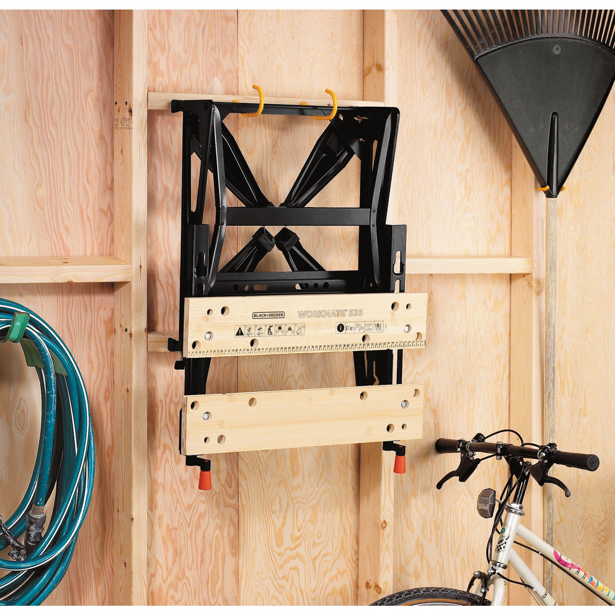 Ein schwarzer Workmate, zusammengeklappt und an einer Wand in einer Garage befestigt. Ein Schlauch und Fahrrad sind ebenfalls sichtbar.