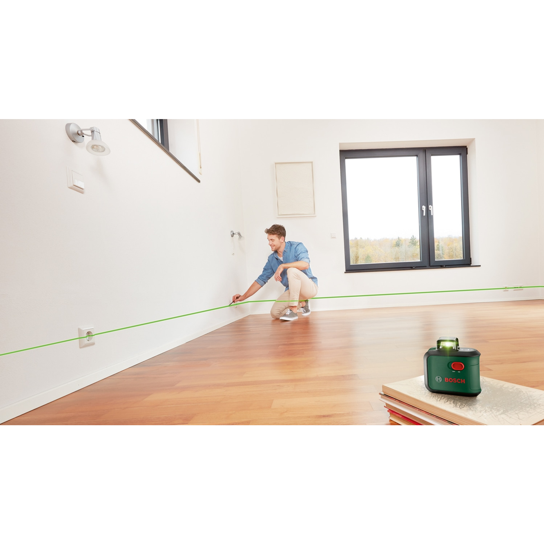 Mężczyzna w pokoju używający zielonego poziomicy laserowej. Poziomica jest na książkach. Drewniana podłoga.
