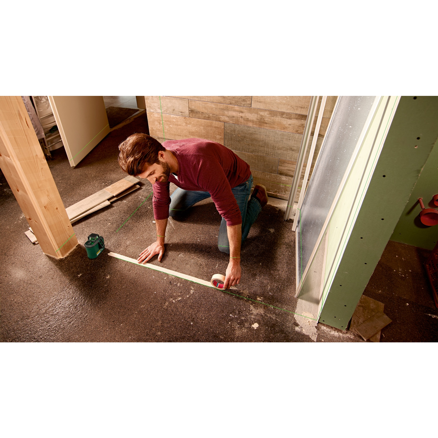 Mężczyzna klęczy, mierząc linię na podłodze. Zielony poziom laserowy, drewniana rama, panel szklany.