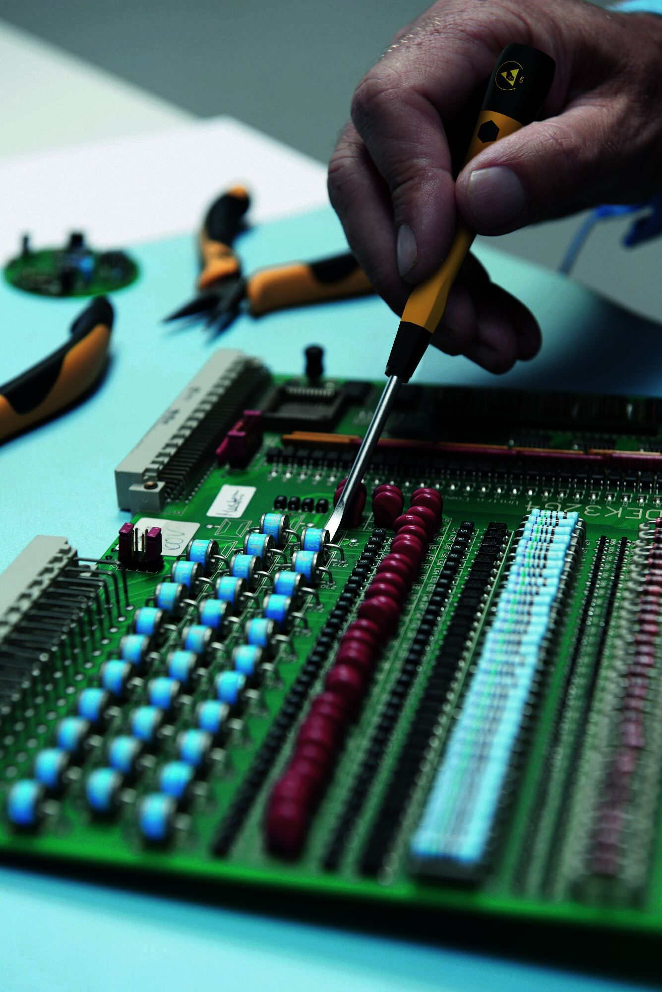 Ręka używa żółtego śrubokręta na zielonej płytce drukowanej z niebieskimi i czerwonymi elementami.