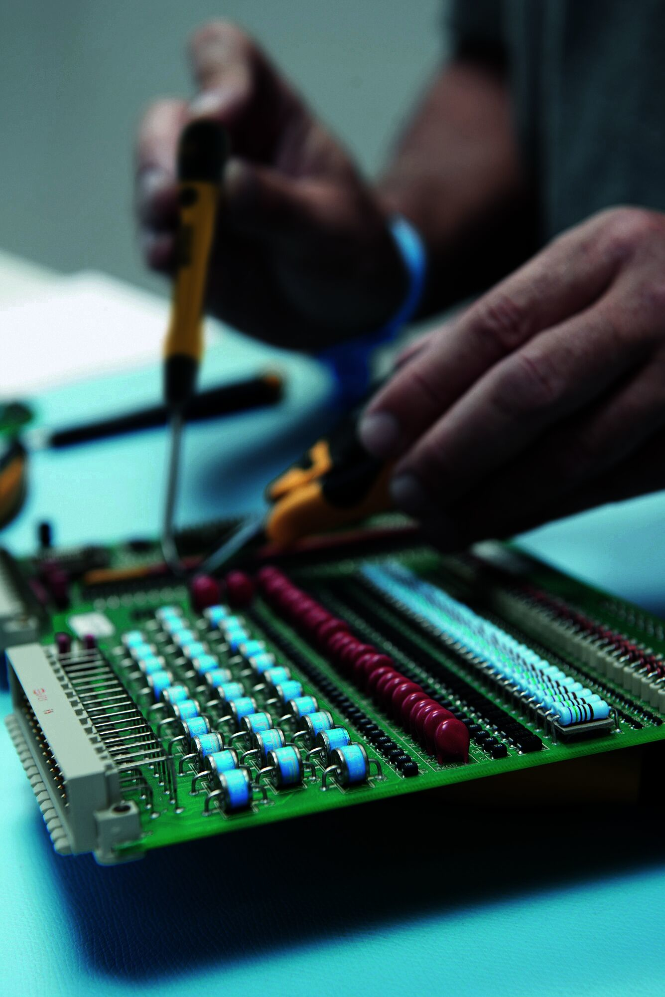 Ręka używa żółtego śrubokręta na zielonej płytce drukowanej z niebieskimi i czerwonymi elementami.