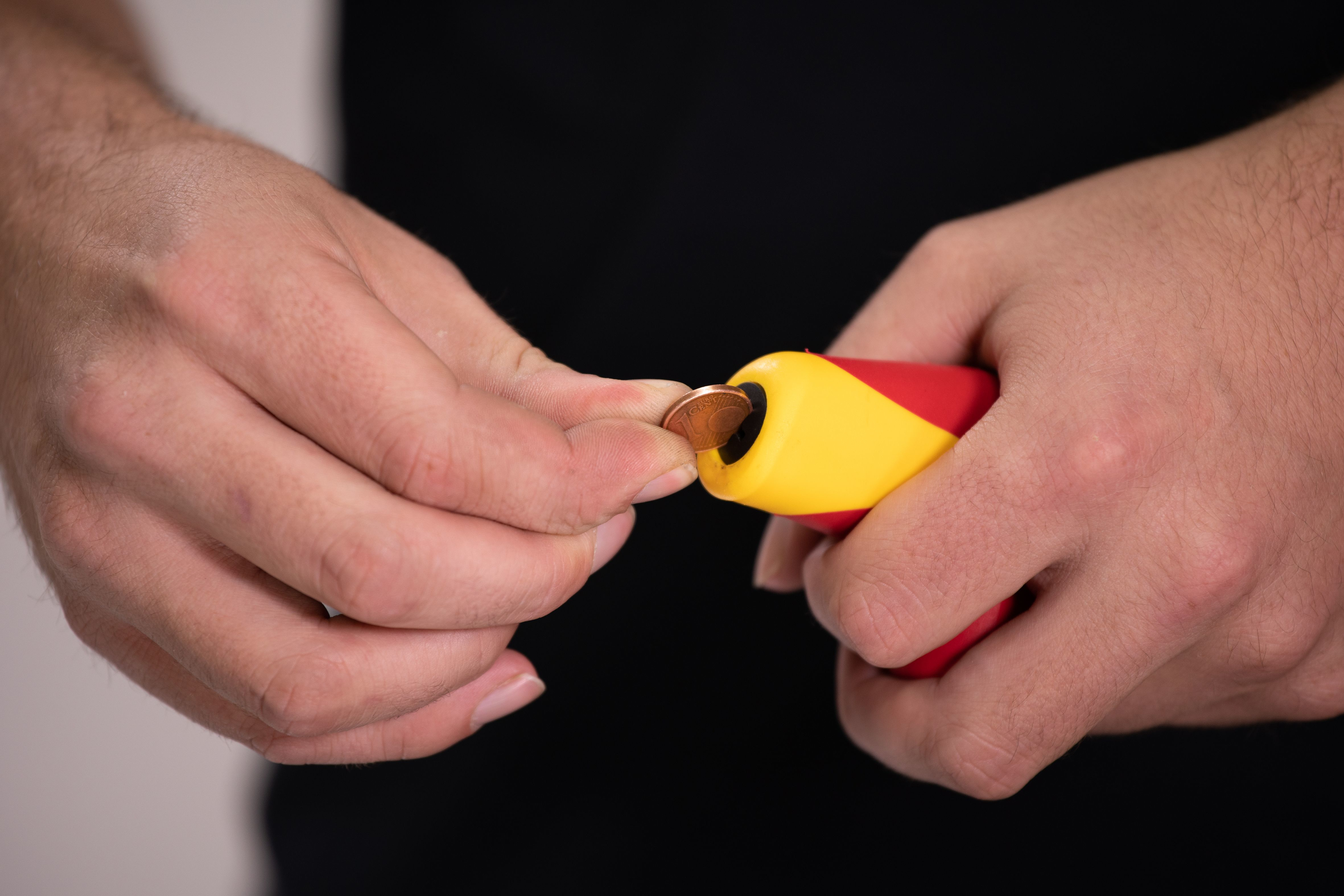 Mani che inseriscono una moneta nella fessura di uno strumento rosso e giallo, primo piano.
