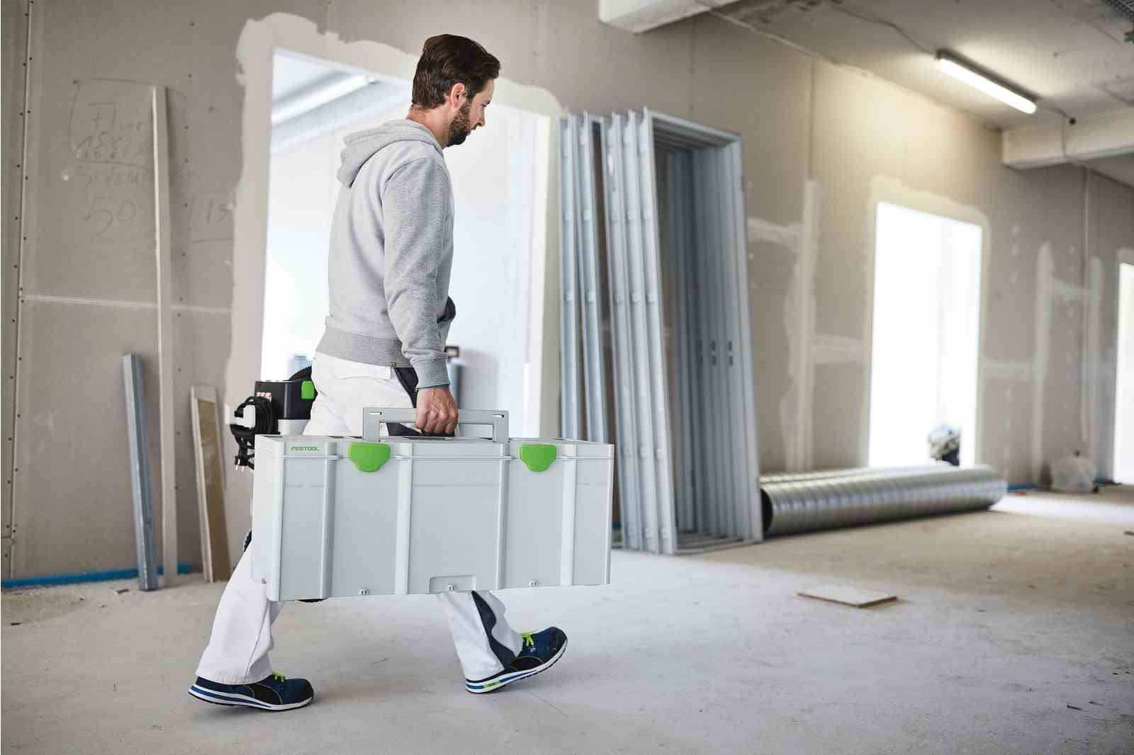 Mann trägt eine weiße Festool-Werkzeugkiste auf einer Baustelle. Arbeitssachen tragend.