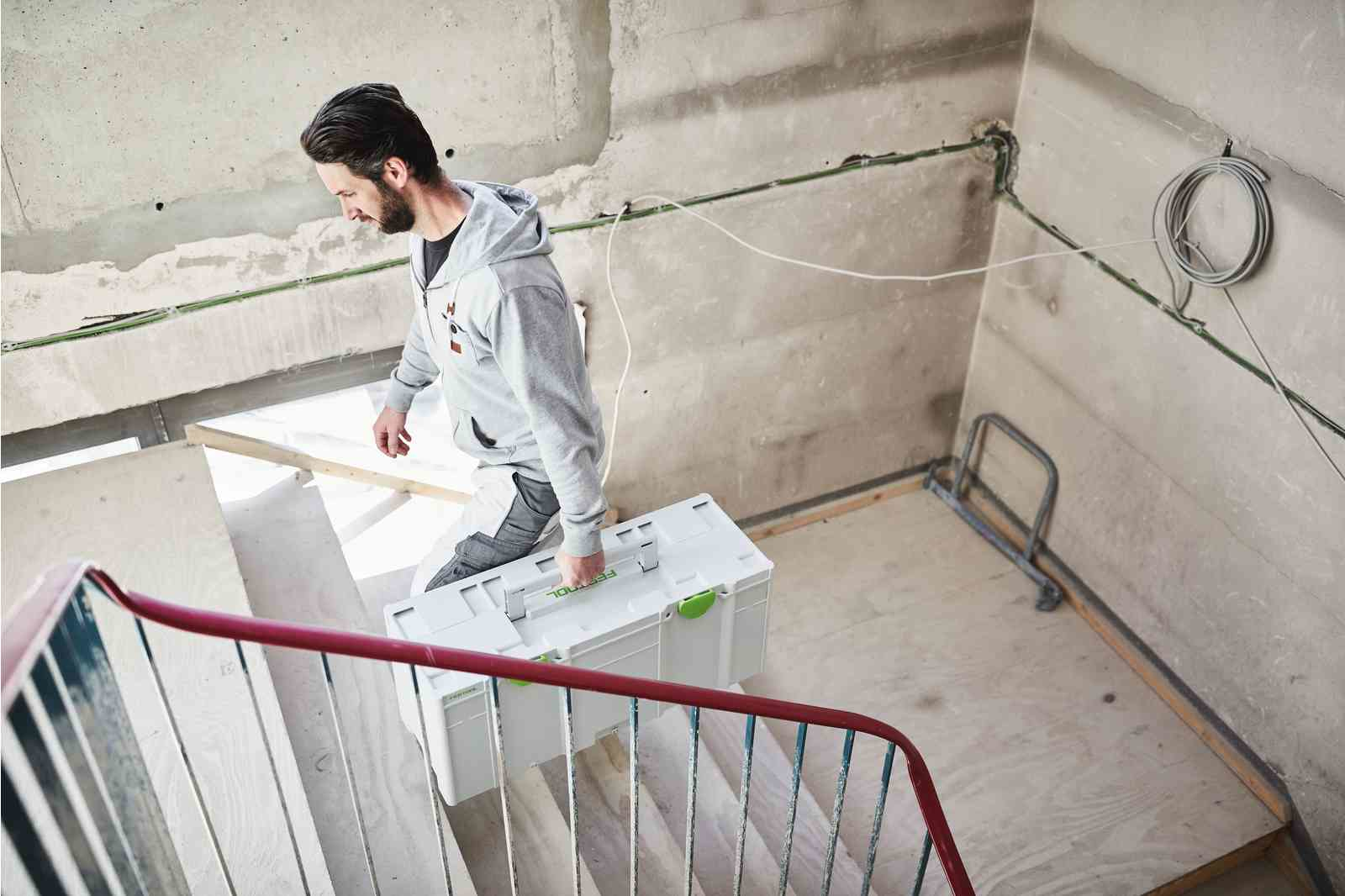 Mann trägt eine weiße Festool-Werkzeugkiste die Treppe hoch. Baustelle im Hintergrund.