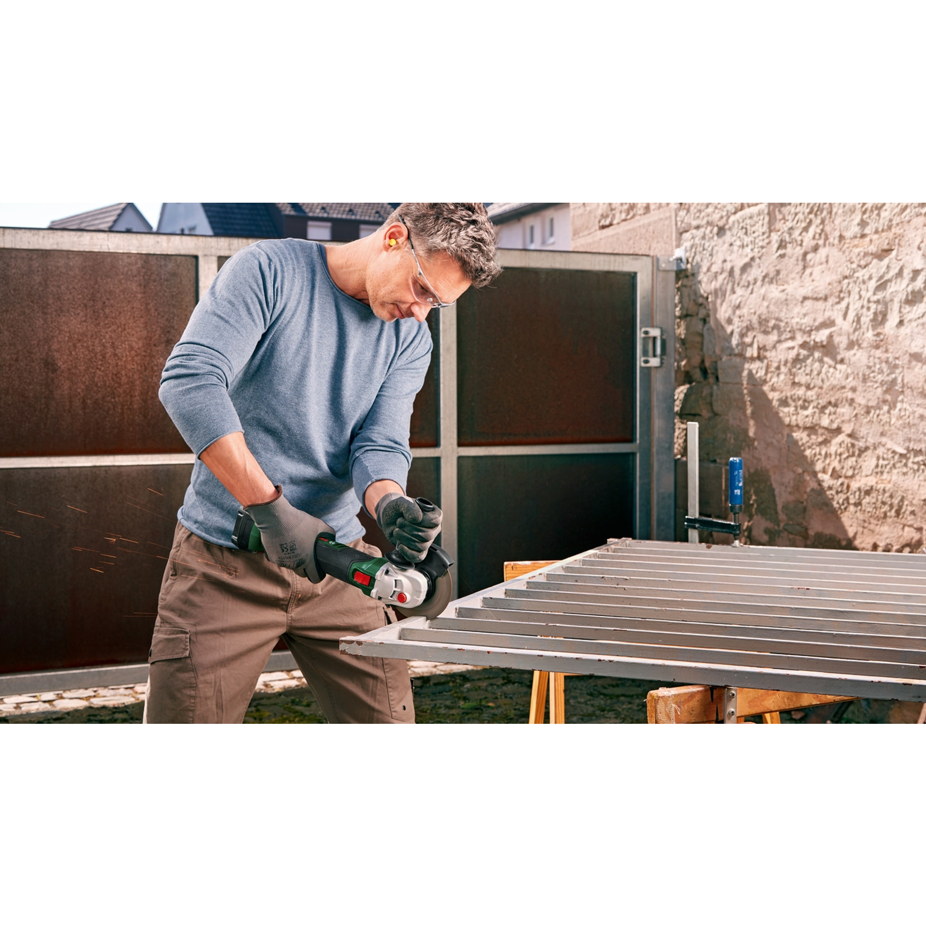 Mężczyzna szlifuje metalową bramę szlifierką. Nosi okulary ochronne. Iskry lecą. Tło: brama, budynek, niebo.