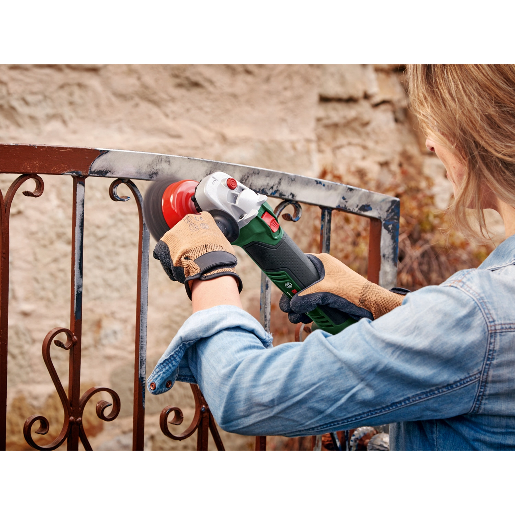 Kobieta używa szlifierki do metalowej balustrady. Iskry lecą. Nosi rękawice. Tło: ceglany mur i balustrada.