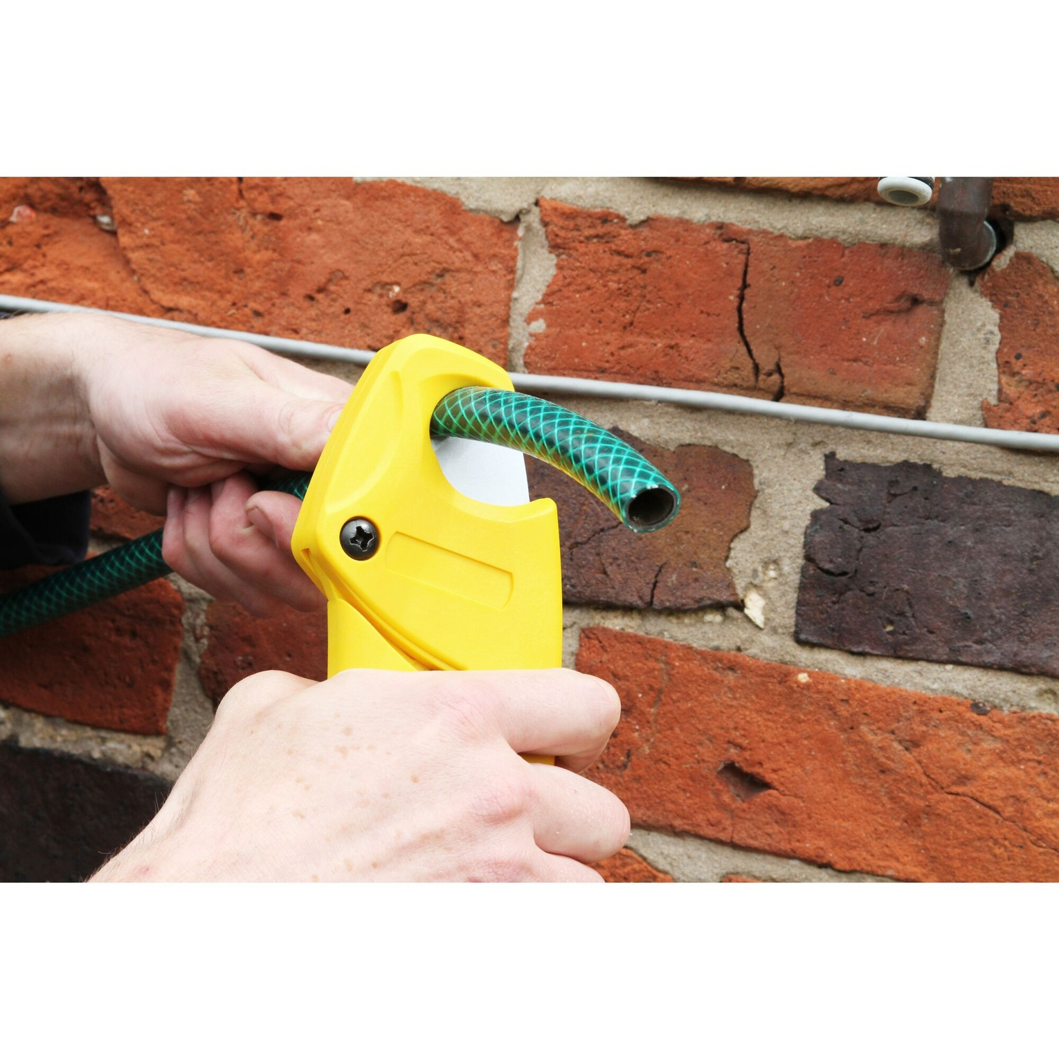 Manos cortando una manguera verde con un cortador amarillo, contra una pared de ladrillos.