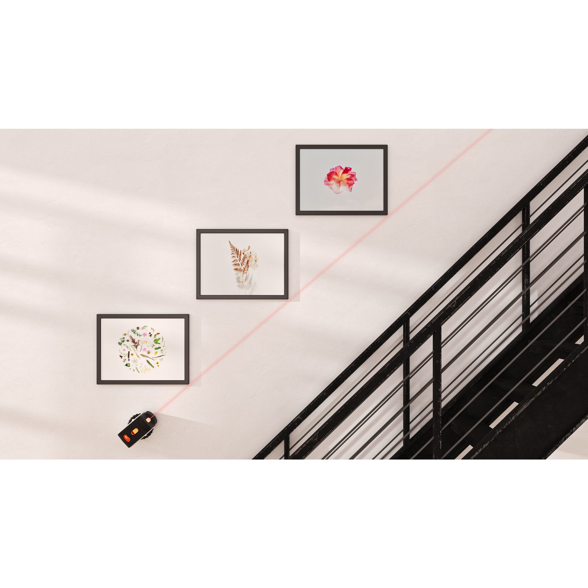 Nivel láser proyecta una línea roja a través de una pared con tres cuadros enmarcados y una escalera.