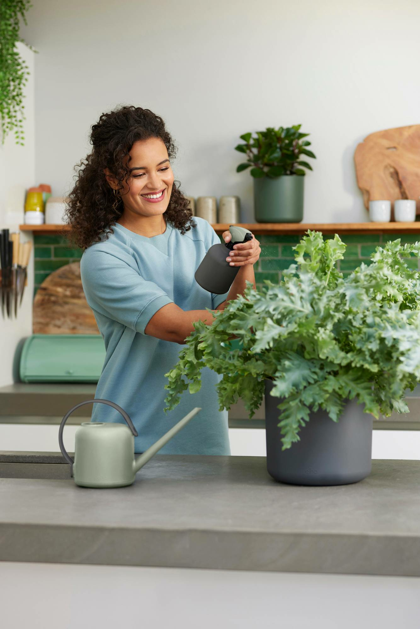 Donna che spruzza una pianta in casa con un flacone spray grigio. Cucina sullo sfondo.