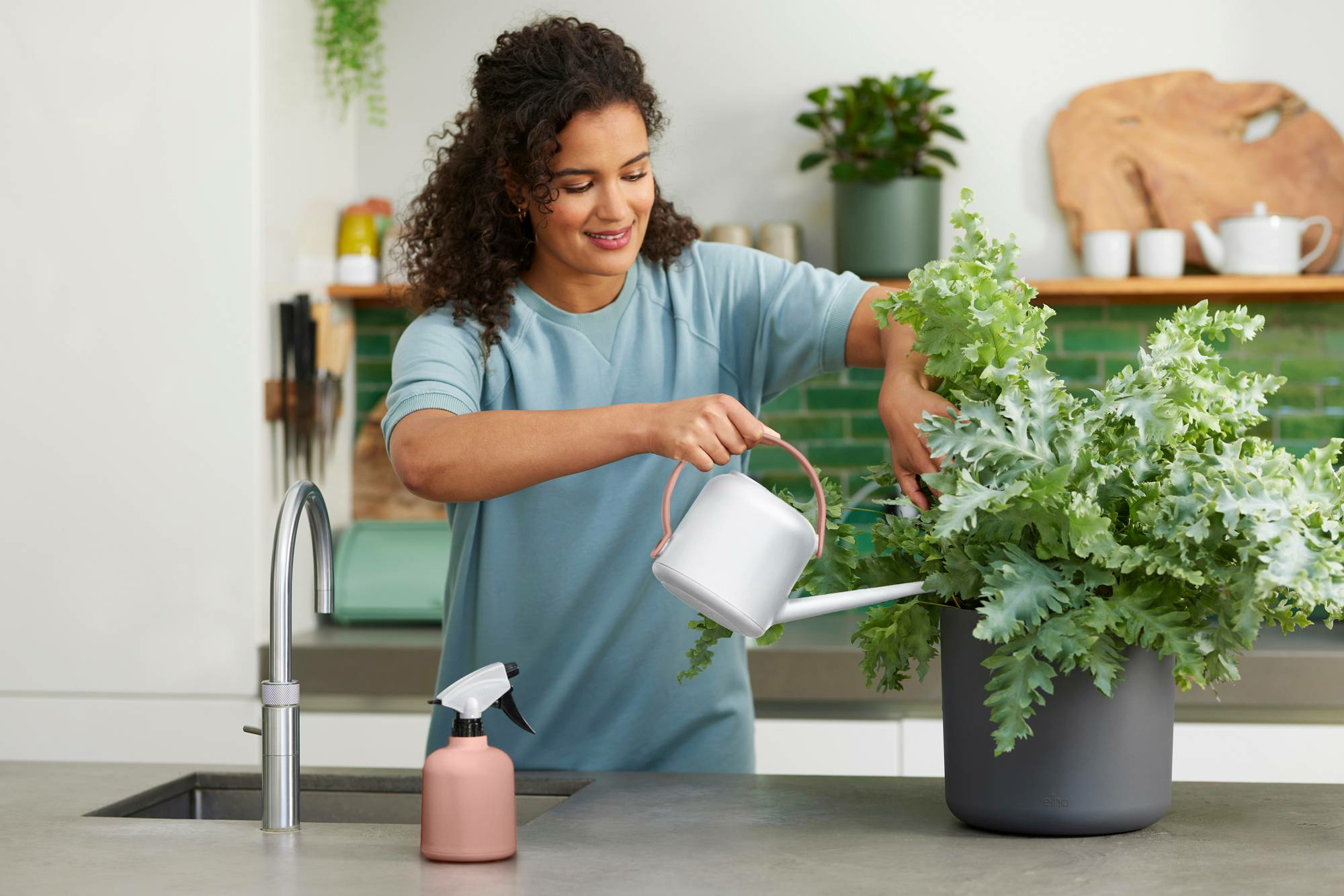 Eine Frau gießt eine Pflanze mit einer Gießkanne und besprüht eine Pflanze mit einem rosa Sprüher in einer Küche.