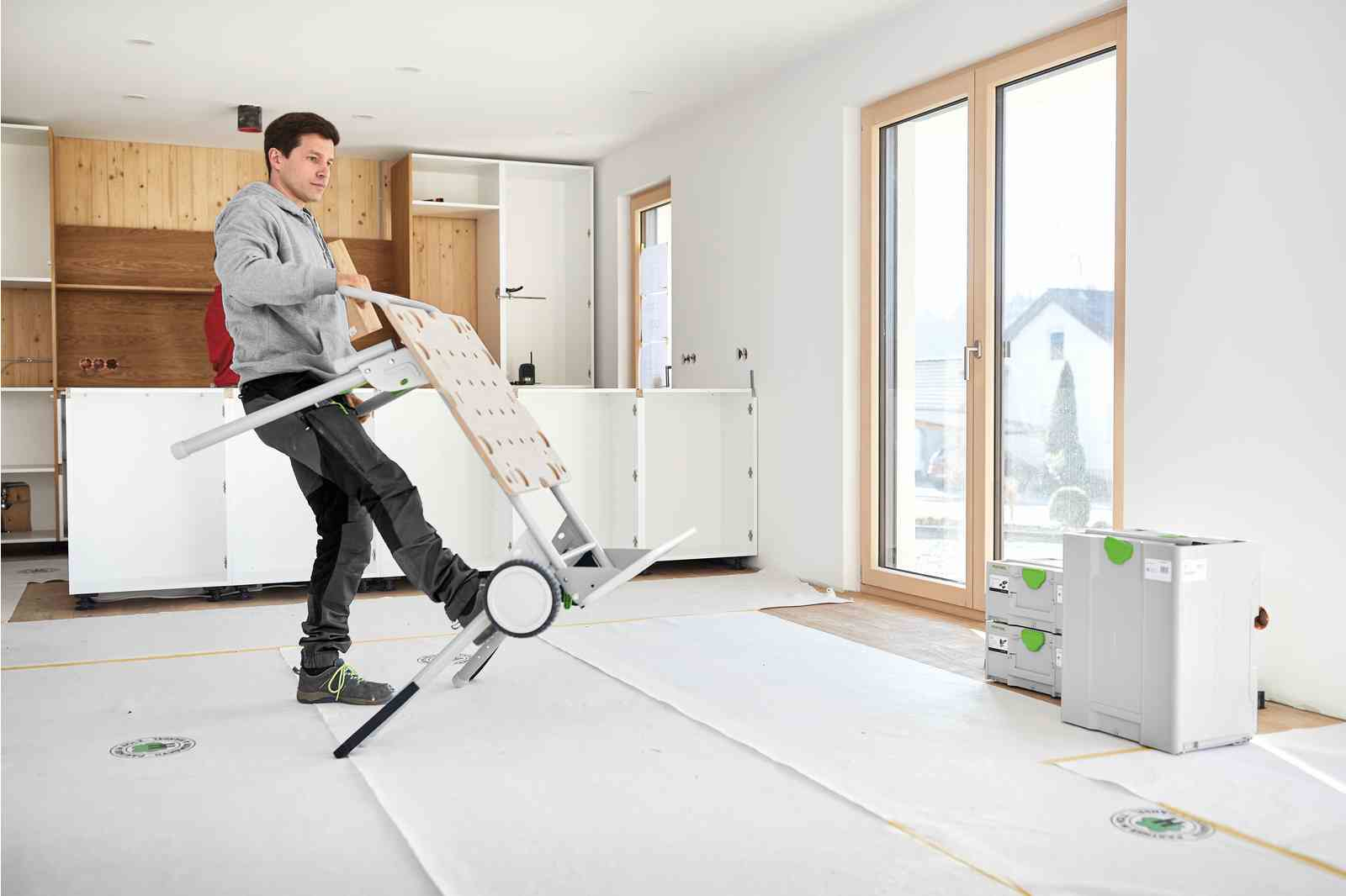 Ein Mann trägt einen faltbaren Arbeitstisch. Der Mann ist in einem Raum mit weißen Wänden und Holzschränken.