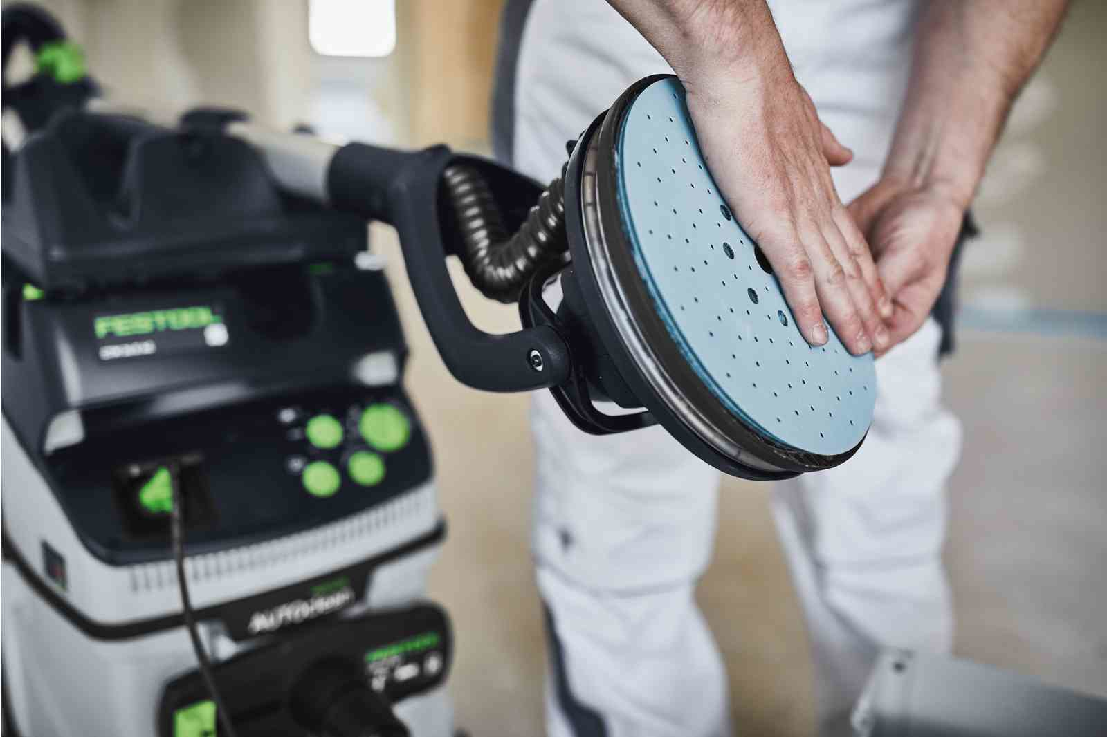 Osoba używająca szlifierki na ścianie. Szlifierka jest podłączona do odkurzacza.