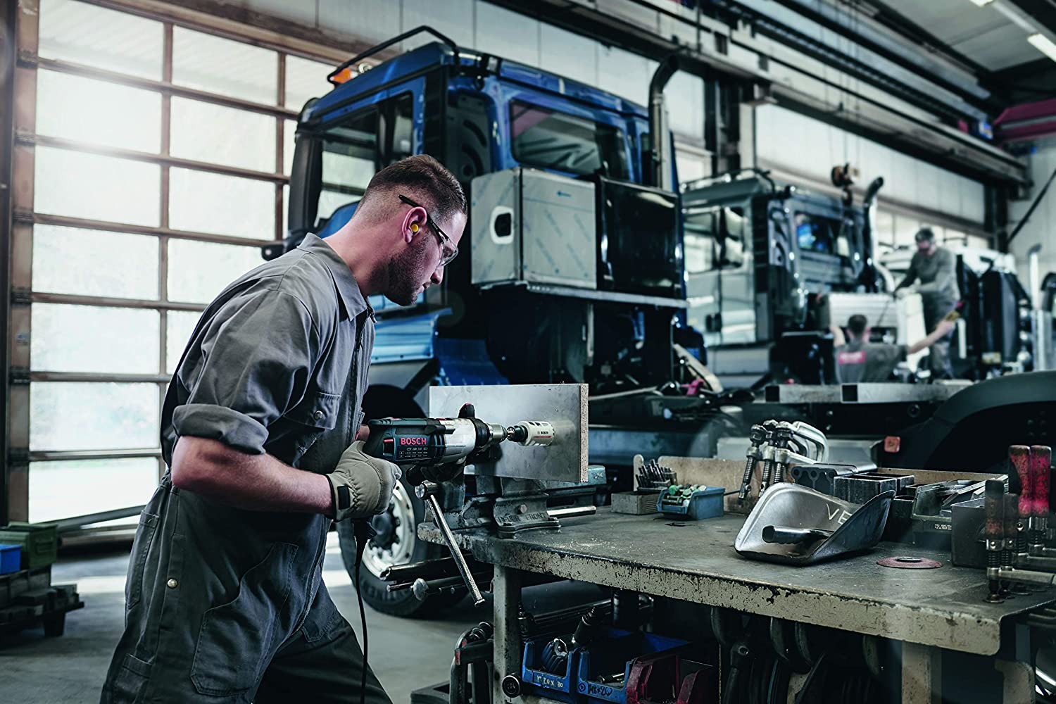 Ein Mann benutzt einen Bohrer auf einem Metallblech, das in einem Schraubstock eingespannt ist. Ein LKW ist im Hintergrund.