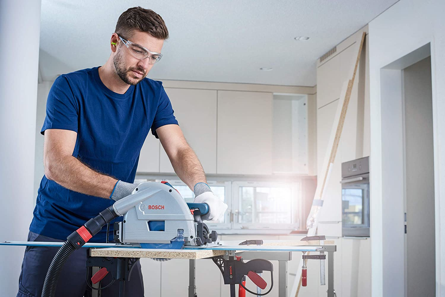 Un uomo con occhiali di sicurezza taglia legno con una sega elettrica in cucina.