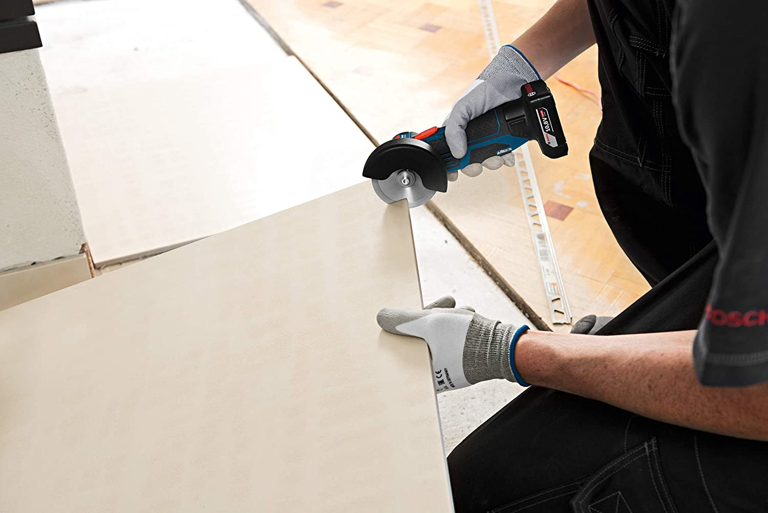 Ein Arbeiter schneidet eine Fliese mit einem Elektrowerkzeug. Das Werkzeug ist blau und schwarz. Der Arbeiter trägt Handschuhe.