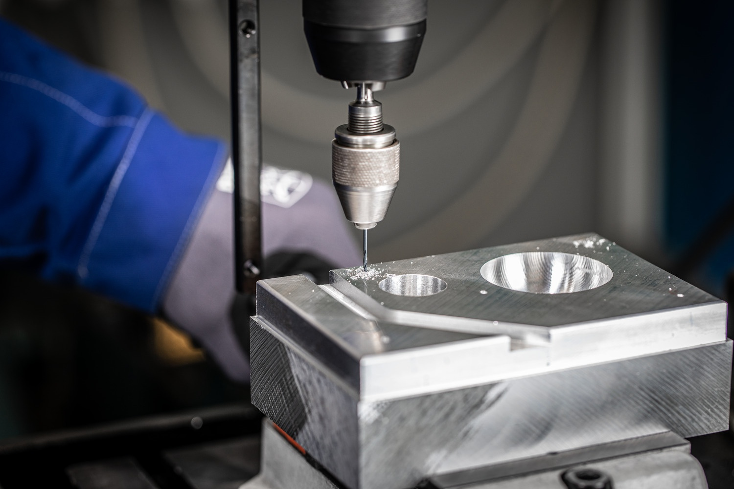 Ein Bohrer durchbohrt einen Metallblock, der von einer Maschine gehalten wird. Eine behandschuhte Hand führt den Prozess.