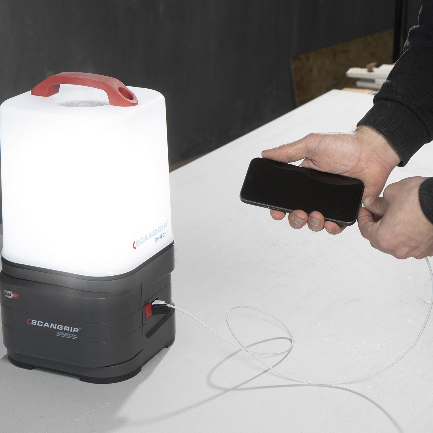 Eine Person verbindet ein Smartphone mit einer weiß-grauen Laterne mit einem USB-Kabel. Der Markenname ist sichtbar.