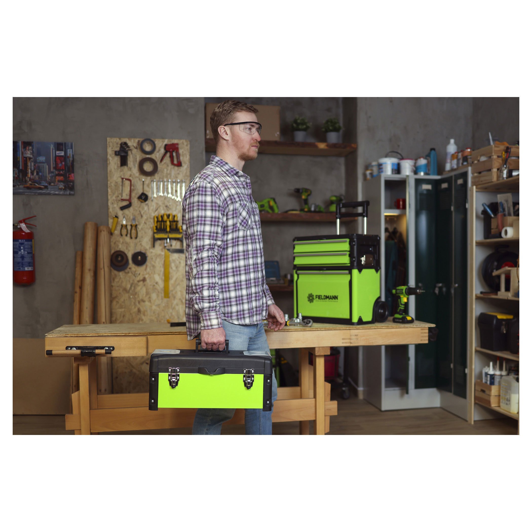 Mężczyzna w koszuli w kratę niesie zieloną skrzynkę narzędziową. Tło warsztatu.