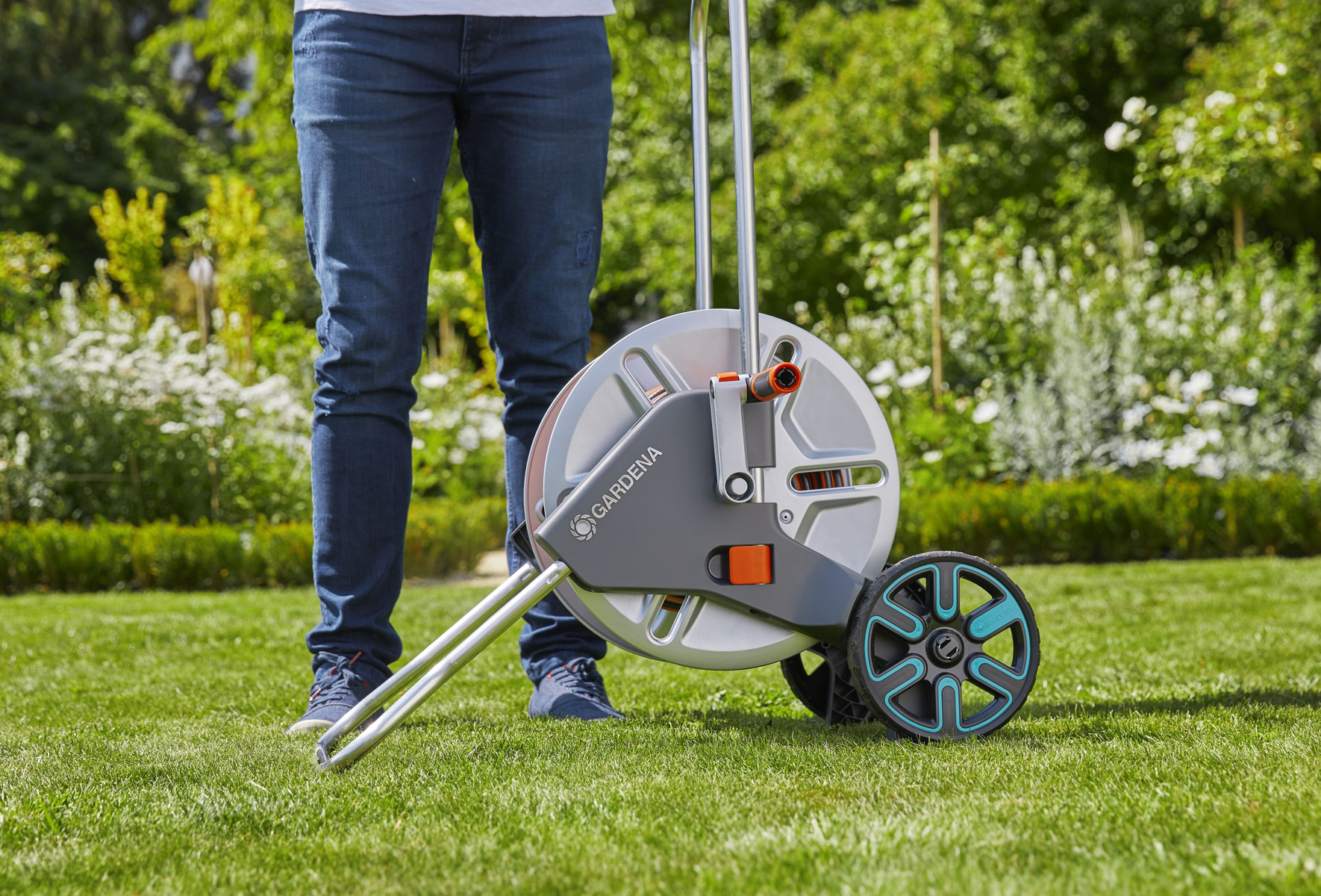 Person steht im Gras und hält eine Schlauchtrommel auf Rädern, im Hintergrund ein Garten.